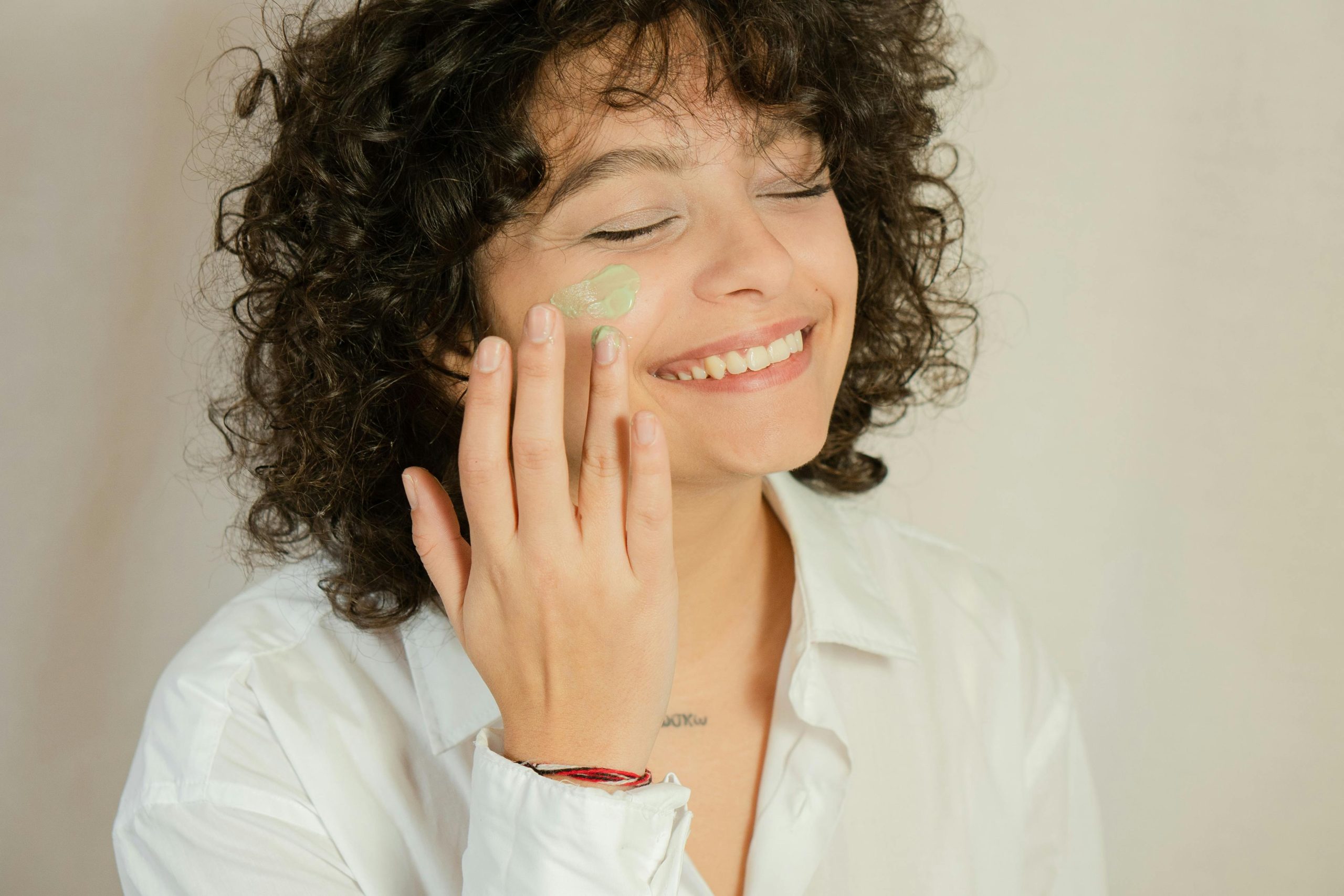 Inicio Close-up of a smiling woman applying facial cream with eyes closed.