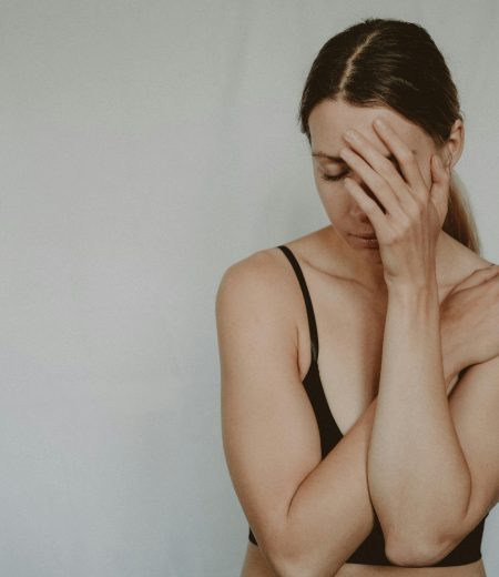 Young slender female with closed eyes covering breast and face on grey background